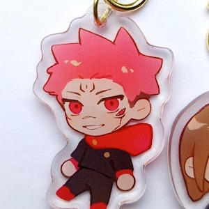 May include: A clear acrylic keychain featuring a cartoon character with red hair, red eyes, and a red scarf. The character is wearing a black uniform with a red collar. The keychain is attached to a gold metal ring.