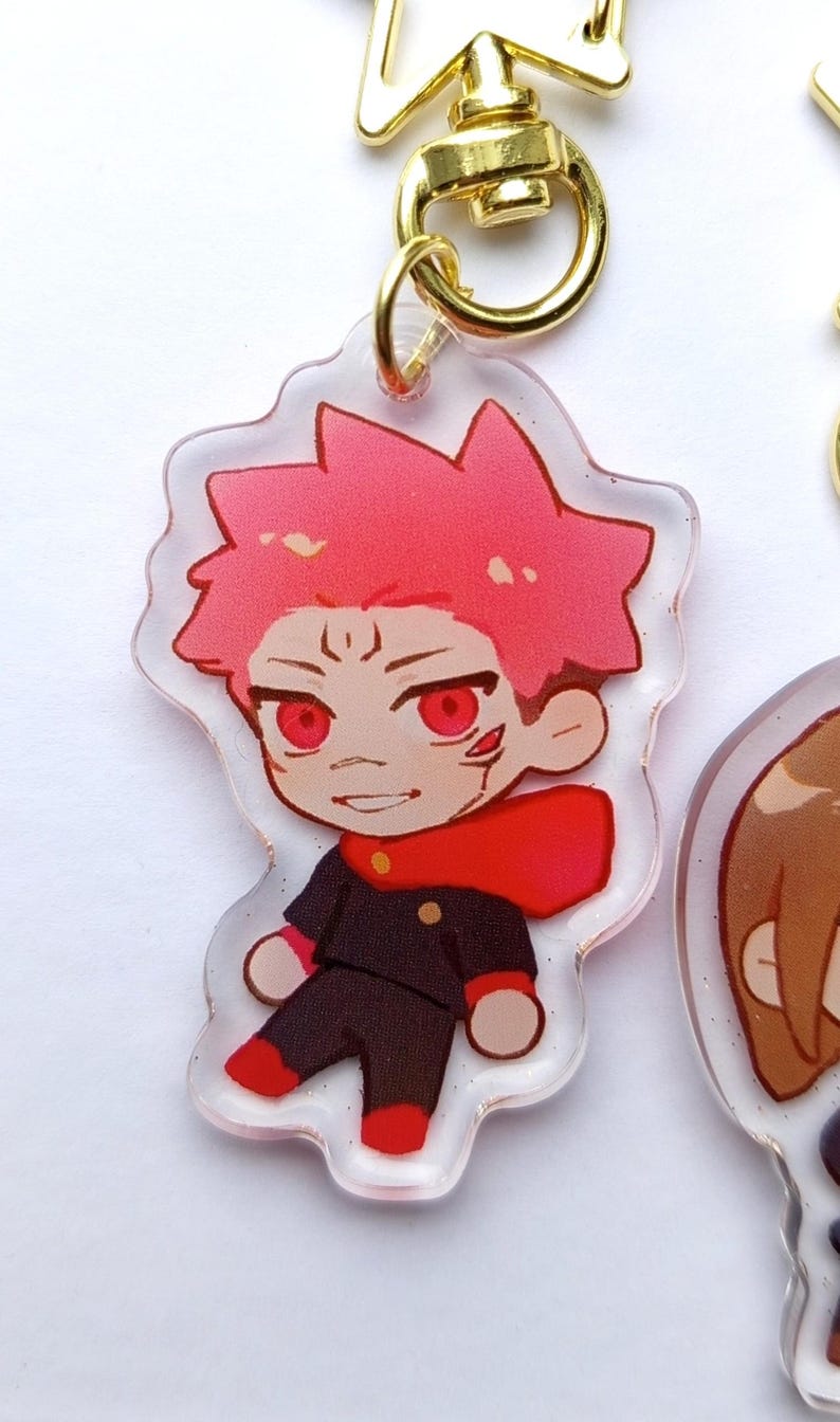 May include: A clear acrylic keychain featuring a cartoon character with red hair, red eyes, and a red scarf. The character is wearing a black uniform with a red collar. The keychain is attached to a gold metal ring.