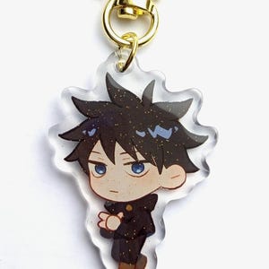 May include: A clear acrylic keychain featuring a cartoon character with black hair, a blue shirt, and a black jacket. The keychain has a gold star-shaped clasp.