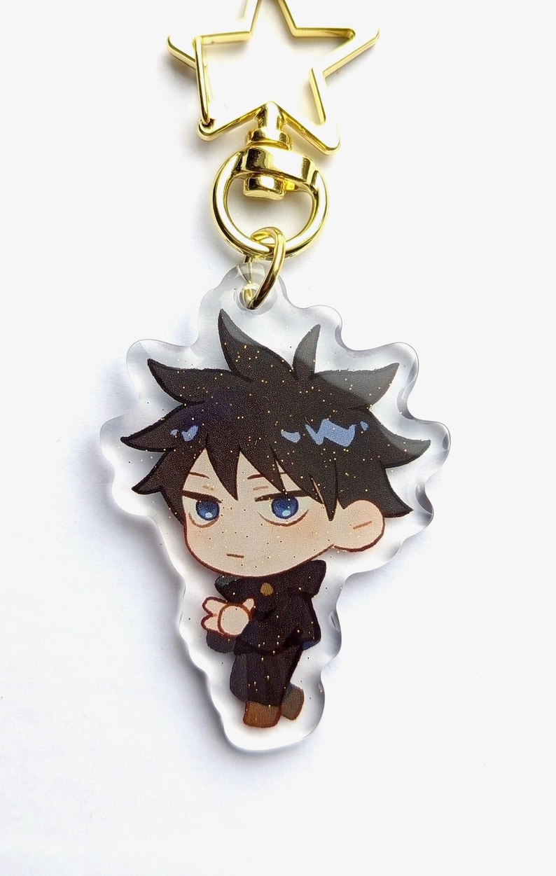 May include: A clear acrylic keychain featuring a cartoon character with black hair, a blue shirt, and a black jacket. The keychain has a gold star-shaped clasp.