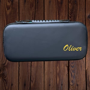 Può includere: Una custodia rigida nera con chiusura a zip e il nome "Oliver" stampato in giallo sulla parte superiore.