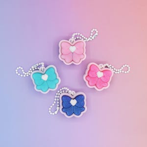 May include: Four colorful bow-shaped keychains in pink, teal, blue, and pink, each with a white heart in the center. The keychains are attached to silver-colored ball chains. The background is a gradient of purple, pink, and orange.