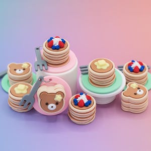 May include: A collection of miniature pancake-themed charms and keychains. The pancakes are stacked with toppings like butter, whipped cream, and berries. Some charms feature a bear design. Gray forks and pink, green, and white platforms are also present.