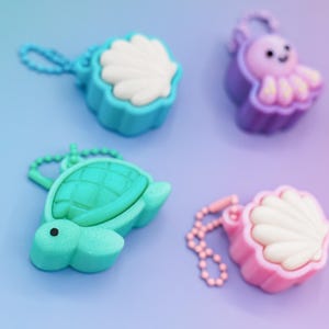 May include: Four colorful keychains in the shape of sea creatures. There is a teal turtle, a purple octopus, and two seashell-shaped keychains in pink and teal. Each keychain has a matching chain.
