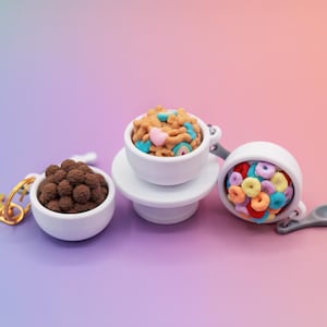 May include: Three miniature cereal bowl keychains. One bowl is filled with chocolate cereal balls, another with colorful cereal pieces, and the third with cereal and marshmallows. The keychains are on a gradient background.