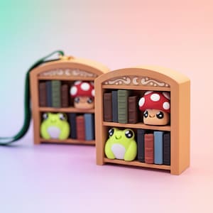 May include: Two miniature wooden bookshelves with arched tops. Each shelf is filled with tiny books and features a smiling mushroom and a frog. The mushroom has a red cap with white spots. The frog is lime green.