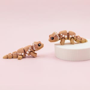 May include: Two brown articulated gecko toys with white spots. One gecko is on a white platform, and the other is on a pink background. The geckos have large, dark eyes and are posed as if interacting with each other.