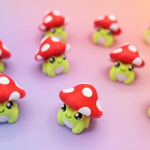 K&ouml;nnte beinhalten: Eine Ansammlung kleiner, skurriler Figuren. Jede Figur zeigt ein hellgr&uuml;nes, froschartiges Wesen mit einem l&auml;chelnden Gesicht, das einen roten Pilzhut mit wei&szlig;en Punkten tr&auml;gt. Der Hintergrund hat einen Farbverlauf von Lila zu Orange.