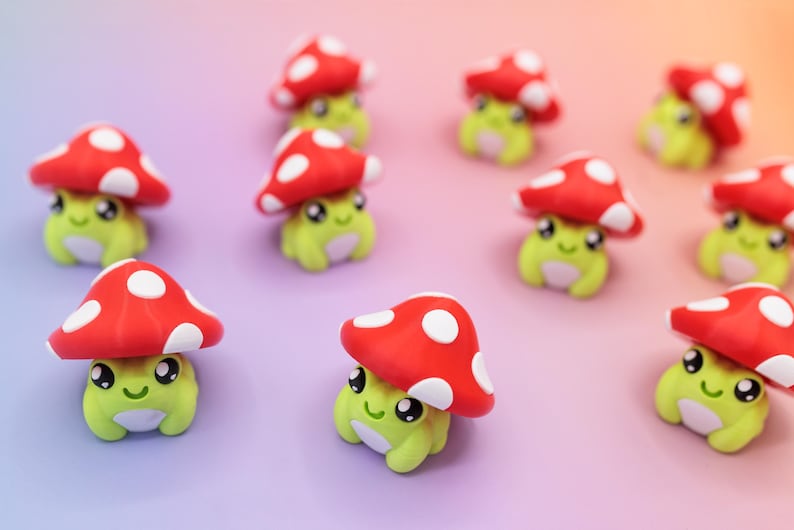 K&ouml;nnte beinhalten: Eine Ansammlung kleiner, skurriler Figuren. Jede Figur zeigt ein hellgr&uuml;nes, froschartiges Wesen mit einem l&auml;chelnden Gesicht, das einen roten Pilzhut mit wei&szlig;en Punkten tr&auml;gt. Der Hintergrund hat einen Farbverlauf von Lila zu Orange.