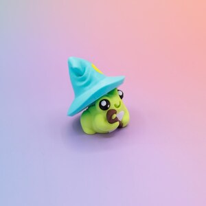 May include: A whimsical, handmade figurine of a green creature wearing a blue wizard hat with a yellow star. The creature has large, expressive eyes and is holding a small brown object. The background is a gradient of purple and orange.