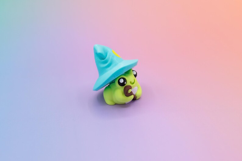 May include: A whimsical, handmade figurine of a green creature wearing a blue wizard hat with a yellow star. The creature has large, expressive eyes and is holding a small brown object. The background is a gradient of purple and orange.