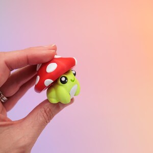 K&ouml;nnte beinhalten: Eine kleine, skurrile Figur eines gr&uuml;nen Frosches mit einem roten Pilzhut. Der Pilz hat wei&szlig;e Punkte. Der Frosch hat gro&szlig;e schwarze Augen und einen l&auml;chelnden Ausdruck. Die Figur wird in einer Hand vor einem Farbverlaufshintergrund gehalten.