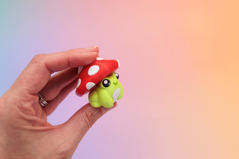 K&ouml;nnte beinhalten: Eine kleine, skurrile Figur eines gr&uuml;nen Frosches mit einem roten Pilzhut. Der Pilz hat wei&szlig;e Punkte. Der Frosch hat gro&szlig;e schwarze Augen und einen l&auml;chelnden Ausdruck. Die Figur wird in einer Hand vor einem Farbverlaufshintergrund gehalten.