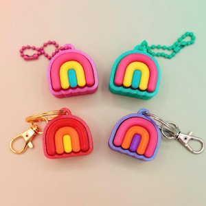 May include: Four colorful rainbow-shaped keychains. Each keychain features a rainbow design with a different color scheme: pink, teal, red, and purple. The keychains have different attachments, including a chain and a clasp.