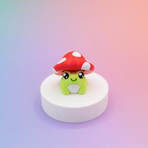 K&ouml;nnte beinhalten: Eine skurrile gr&uuml;ne Froschfigur mit einem roten Pilzhut mit wei&szlig;en Punkten. Der Frosch sitzt auf einem wei&szlig;en, zylindrischen Sockel vor einem Hintergrund aus Blau, Rosa und Orange.