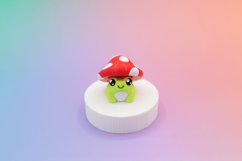 K&ouml;nnte beinhalten: Eine skurrile gr&uuml;ne Froschfigur mit einem roten Pilzhut mit wei&szlig;en Punkten. Der Frosch sitzt auf einem wei&szlig;en, zylindrischen Sockel vor einem Hintergrund aus Blau, Rosa und Orange.