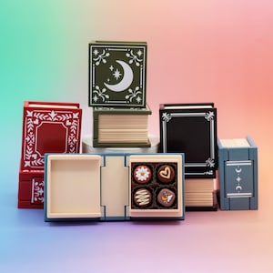 May include: A collection of decorative book-shaped boxes in various colors, including red, green, black, and blue. One box is open, revealing four chocolates with different designs. The boxes feature decorative patterns and celestial motifs.