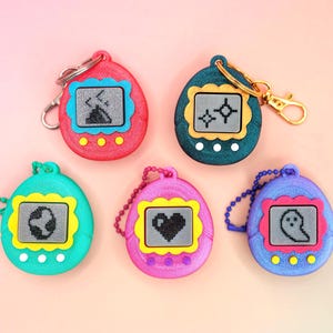 May include: A collection of five colorful, retro handheld digital pet toys. Each toy features a small screen displaying a pixelated graphic, with a different icon on each. The toys are in shades of red, teal, pink, blue, and green.