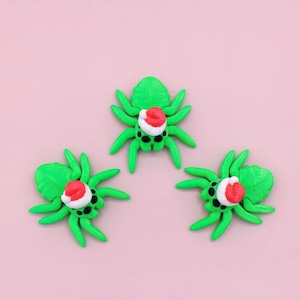 May include: Three green spiders with eight legs each, wearing small red and white Santa hats, are arranged on a pink background. The spiders have black eyes and are made of a smooth material.