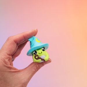 May include: A whimsical, green, wizard-themed figurine with a blue hat and a yellow crescent moon. The figurine is holding a brown staff and has large, expressive eyes. The background is a gradient of pink, blue, and orange.