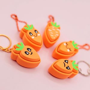 May include: A collection of orange carrot-shaped keychains with green tops and smiling faces. Some keychains have gold-colored clasps, while others have silver-colored clasps. The keychains are arranged on a light pink surface.