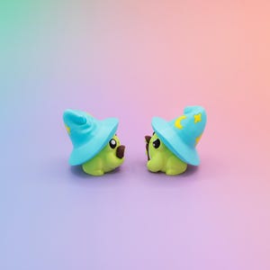 May include: Two small, green, cartoon-like figures wearing light blue wizard hats with yellow crescent moons and stars. The figures have brown noses and are set against a gradient background of purple, pink, and green.