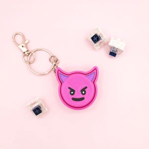 May include: A pink devil face keychain with a silver clasp and ring. The devil face has a black angry expression and purple accents. Two clear mechanical keyboard switches are also in the image.