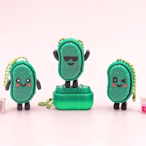 May include: Three green pickle-shaped keychains with smiling faces and black arms and legs. One keychain is standing on a green base, wearing sunglasses. Each keychain has a small chain and a clear, pink and white cube.