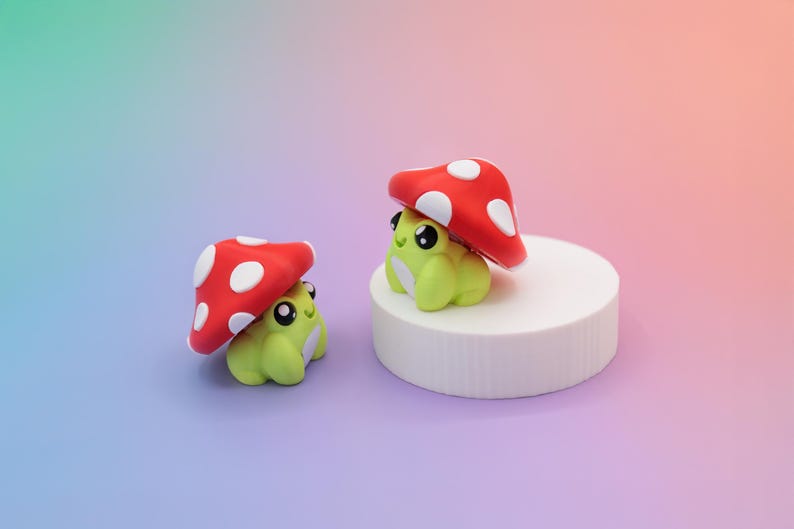 K&ouml;nnte beinhalten: Zwei handgefertigte Froschfiguren, jede mit einem roten Pilzhut mit wei&szlig;en Punkten. Ein Frosch sitzt auf einer wei&szlig;en zylindrischen Plattform. Die Fr&ouml;sche sind hellgr&uuml;n mit schwarzen Augen und wei&szlig;en Akzenten.