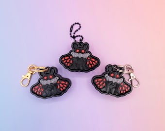 May include: Three black moth-shaped keychains with red and white accents. Each keychain has a different clasp: gold, silver, and a black chain. The keychains are on a gradient purple and pink background.