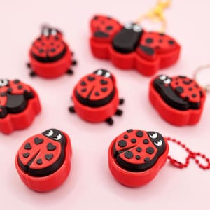May include: A collection of red and black ladybug-shaped charms and keychains. The ladybugs have black spots and eyes, with some featuring heart-shaped markings. The items are arranged on a light pink surface.