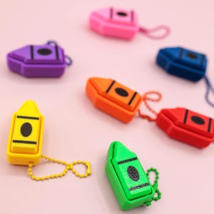 May include: A collection of colorful crayon-shaped keychains in various colors, including yellow, green, orange, red, blue, purple, and pink. Each keychain features a black circle and a small chain for attachment. The keychains are arranged on a light pink surface.