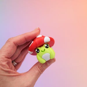 K&ouml;nnte beinhalten: Eine kleine, skurrile Figur einer Frosch mit einem roten Pilzhut mit wei&szlig;en Punkten. Der Frosch ist lindgr&uuml;n mit einem wei&szlig;en Bauch und gro&szlig;en, ausdrucksstarken Augen. Die Figur wird in einer Hand vor einem weichen, farbenfrohen Hintergrund gehalten.