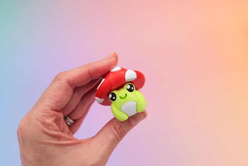 K&ouml;nnte beinhalten: Eine kleine, skurrile Figur einer Frosch mit einem roten Pilzhut mit wei&szlig;en Punkten. Der Frosch ist lindgr&uuml;n mit einem wei&szlig;en Bauch und gro&szlig;en, ausdrucksstarken Augen. Die Figur wird in einer Hand vor einem weichen, farbenfrohen Hintergrund gehalten.