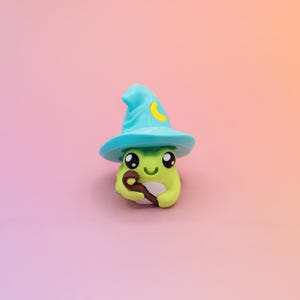May include: A whimsical, lime green frog figurine wearing a turquoise wizard hat with a yellow crescent moon. The frog holds a brown wand and has large, expressive eyes. The background is a gradient of pink and orange.