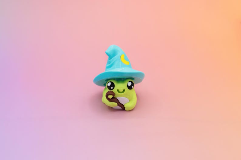 May include: A whimsical, lime green frog figurine wearing a turquoise wizard hat with a yellow crescent moon. The frog holds a brown wand and has large, expressive eyes. The background is a gradient of pink and orange.