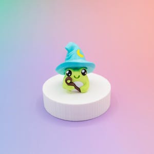 May include: A whimsical, handcrafted frog figurine, wearing a turquoise wizard hat with a yellow crescent moon. The frog holds a brown staff and sits on a white, cylindrical pedestal against a gradient background.