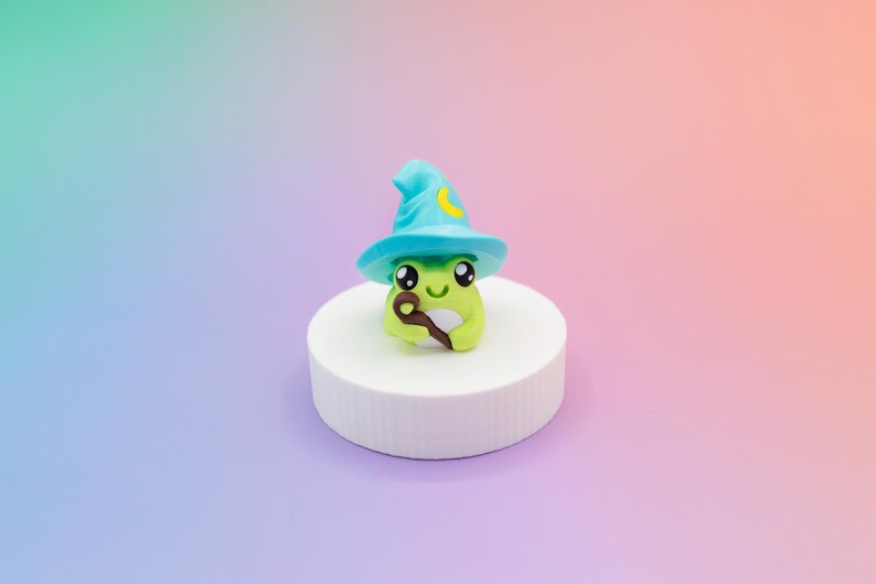 May include: A whimsical, handcrafted frog figurine, wearing a turquoise wizard hat with a yellow crescent moon. The frog holds a brown staff and sits on a white, cylindrical pedestal against a gradient background.