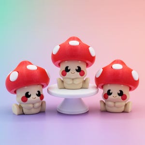 May include: Three whimsical mushroom figurines with red caps and white spots. The mushrooms have smiling faces and are in a sitting position. One mushroom is on a small white pedestal.