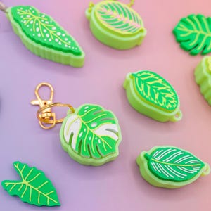 May include: A collection of green leaf-shaped keychains and charms. The keychains feature various leaf designs with white and green details. One keychain has a gold-colored clasp. The items are arranged on a purple and pink background.