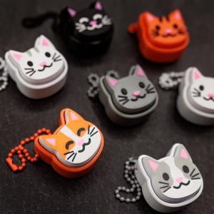 May include: A collection of cat-shaped keychains in various colors, including white, black, orange, and gray. Each keychain features a smiling cat face with pink accents and a small chain for attachment. The keychains are made of a rubbery material.