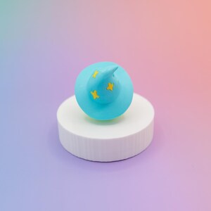 May include: A light blue wizard hat with yellow star accents sits atop a white cylindrical pedestal. The hat has a pointed tip and is set against a gradient background of blue, purple, and orange.