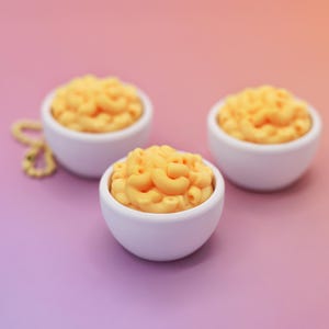 May include: Three small white bowls filled with macaroni and cheese, arranged on a gradient purple and orange background. The macaroni is a bright yellow-orange color. A gold-colored beaded necklace is in the background.