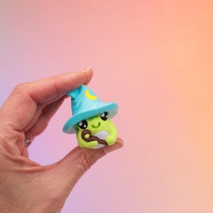 May include: A whimsical, green avocado-shaped figurine wearing a light blue wizard hat with a yellow crescent moon. The figurine has large, expressive eyes and a small, brown staff. The background is a gradient of pink, purple, and orange.