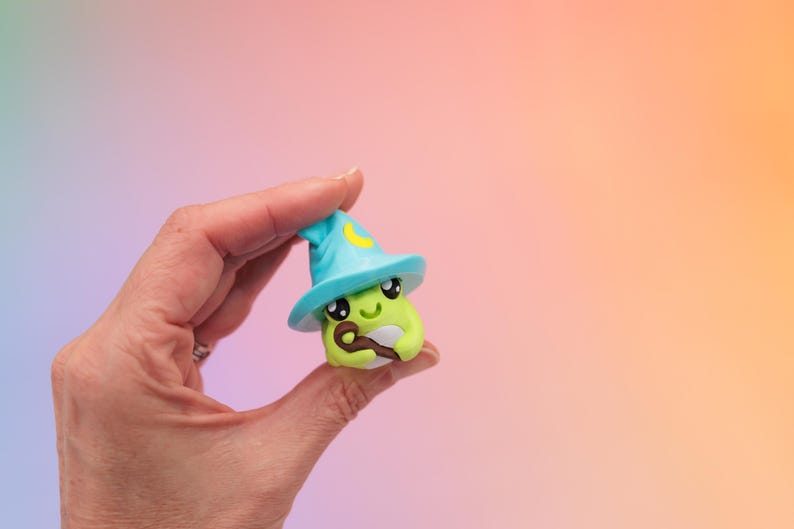 May include: A whimsical, green avocado-shaped figurine wearing a light blue wizard hat with a yellow crescent moon. The figurine has large, expressive eyes and a small, brown staff. The background is a gradient of pink, purple, and orange.