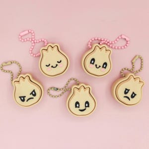 May include: Five dumpling-shaped keychains with different facial expressions. Each keychain is beige with a light brown outline and a metal chain. The keychains have pink and silver-colored chains. The background is a solid pink.