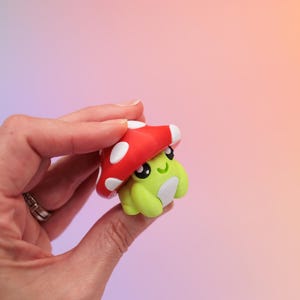 K&ouml;nnte beinhalten: Kleine, skurrile Figur eines gr&uuml;nen Frosches mit rotem Pilzhut mit wei&szlig;en Punkten. Der Frosch hat gro&szlig;e schwarze Augen und einen l&auml;chelnden Ausdruck. Der Hintergrund ist ein Farbverlauf aus Lila, Rosa und Orange.