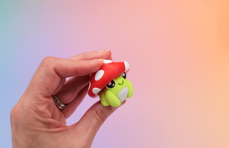 K&ouml;nnte beinhalten: Kleine, skurrile Figur eines gr&uuml;nen Frosches mit rotem Pilzhut mit wei&szlig;en Punkten. Der Frosch hat gro&szlig;e schwarze Augen und einen l&auml;chelnden Ausdruck. Der Hintergrund ist ein Farbverlauf aus Lila, Rosa und Orange.