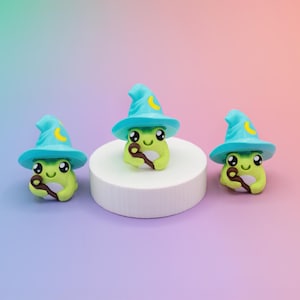 May include: Three whimsical frog figurines, each wearing a turquoise wizard hat with a yellow crescent moon. The frogs are light green and hold a brown wand. They are arranged on a white platform against a gradient background.