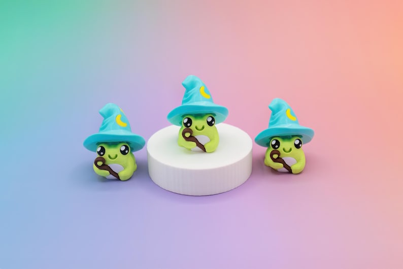 May include: Three whimsical frog figurines, each wearing a turquoise wizard hat with a yellow crescent moon. The frogs are light green and hold a brown wand. They are arranged on a white platform against a gradient background.